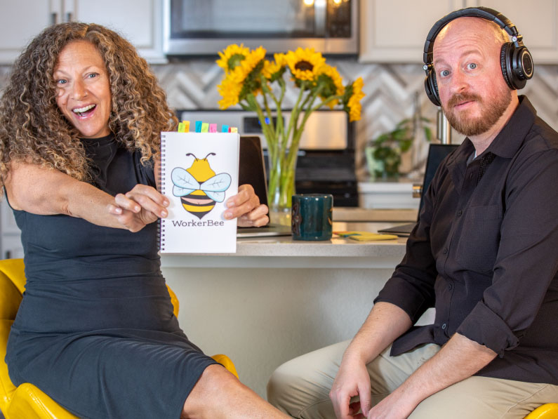 A smiling woman in a black dress holds up a "WorkerBee" notebook with a bee illustration, while a man with headphones sits beside her in a kitchen with sunflowers on the counter in the background.